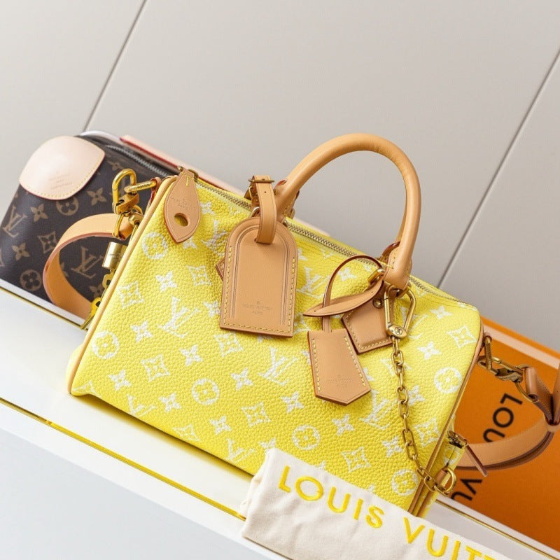 A yellow Louis Vuitton Speedy bag with white monogram print, featuring tan leather handles and tags, displayed on a white shelf next to a Louis Vuitton dust bag and another monogram bag.