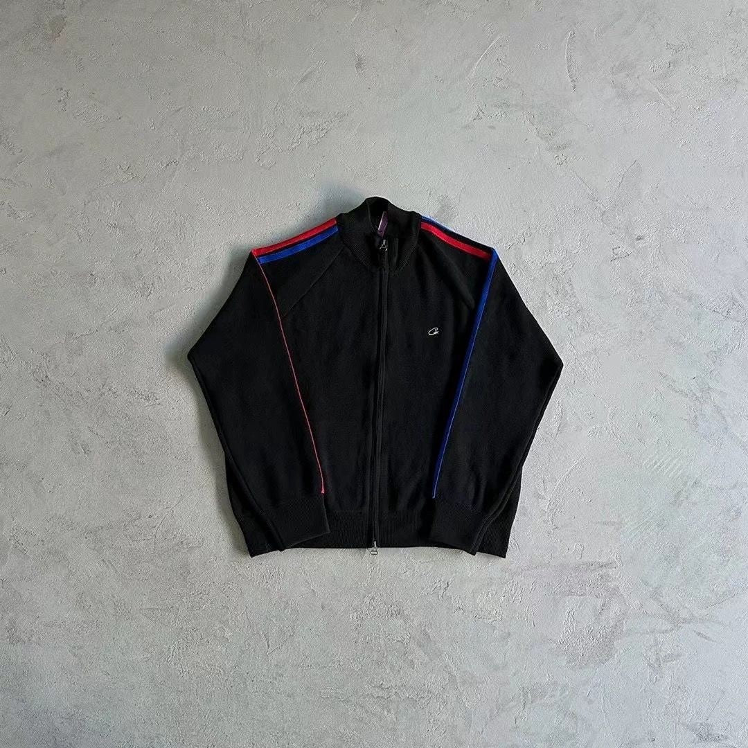 A black zip-up track jacket laid flat on a textured light gray concrete surface. The jacket features red and blue stripes along the shoulders and down the sleeves, with a small white embroidered logo on the left chest.