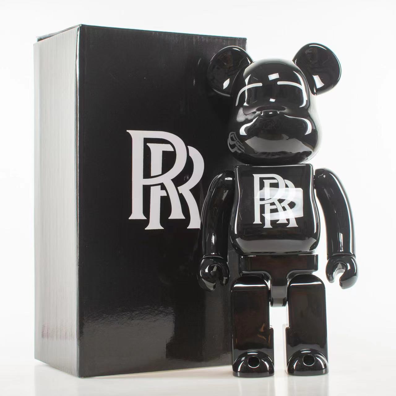 A glossy black bear-shaped collectible figure with a stylized white logo on its chest stands in front of a tall black box featuring a large white monogram.