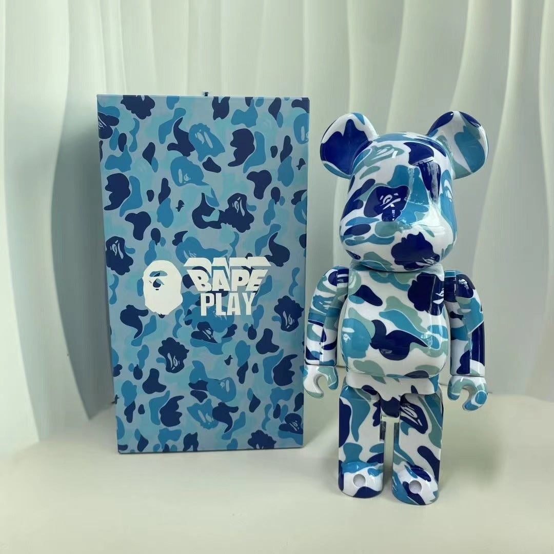A blue and white camouflage Bearbrick figure standing next to its matching branded box, featuring the A Bathing Ape logo and BAPE PLAY text on a light draped background.