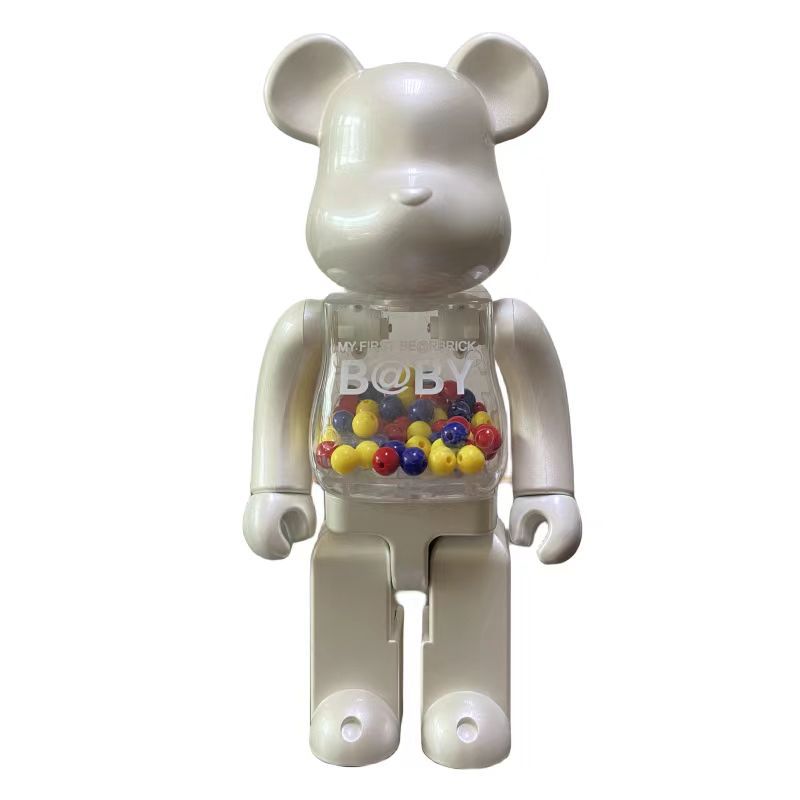 A Bearbrick-style collectible figure in a glossy off-white color, featuring a transparent chest compartment filled with small multicolored round beads and printed with the text 'MY FIRST BEARBRICK' and 'B@Y'.