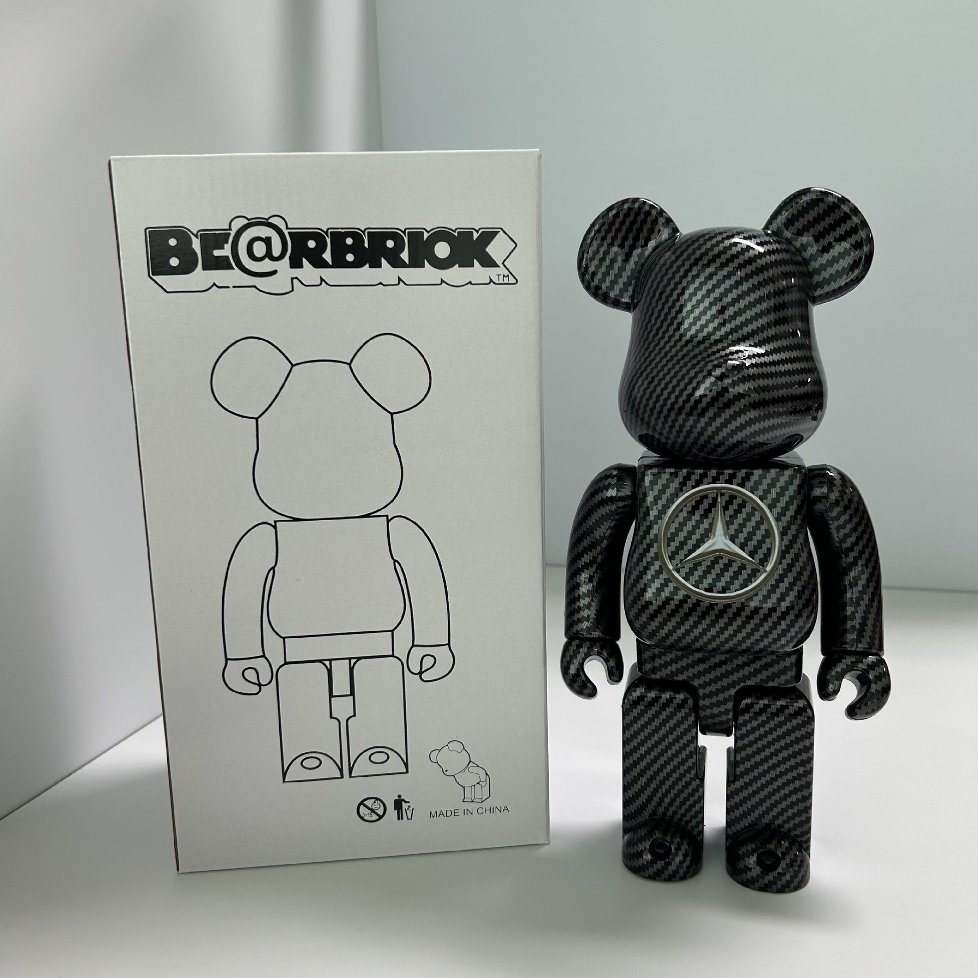 A Bearbrick collectible figure with a black carbon fiber pattern stands next to its white packaging box, which features the BE@RBRIOK logo and a line drawing of the bear figure.