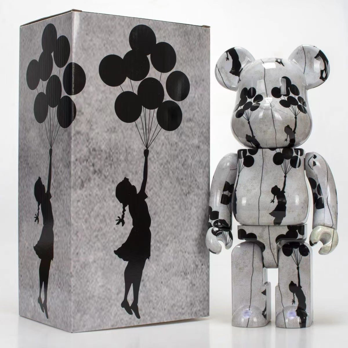 A glossy bear-shaped collectible figure stands beside its matching box. The figure and packaging are decorated with a gray marbled pattern and black silhouettes of a girl holding a cluster of balloons, against a clean white background.