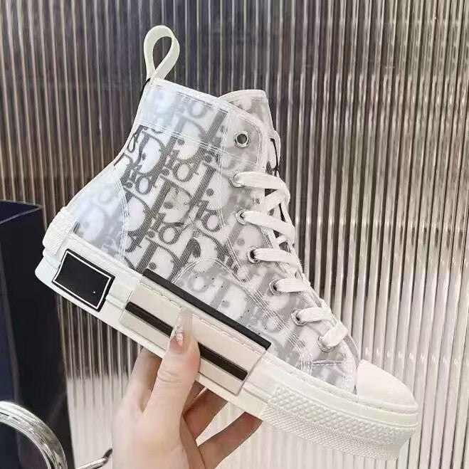 A hand holding a high-top sneaker with a white and gray Dior monogram canvas upper, white laces, and a chunky white sole, set against a metallic ribbed background.