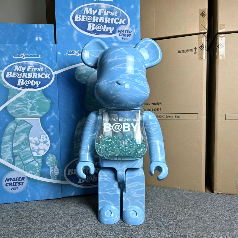 A blue water-marbled Bearbrick Baby collectible figure standing in front of its blue packaging boxes and cardboard shipping cartons on a gray floor.