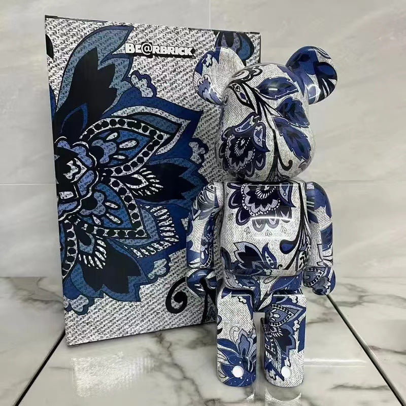 A Bearbrick collectible figure with a blue, white, and black floral mandala pattern stands in front of its matching patterned packaging box on a light marble surface.