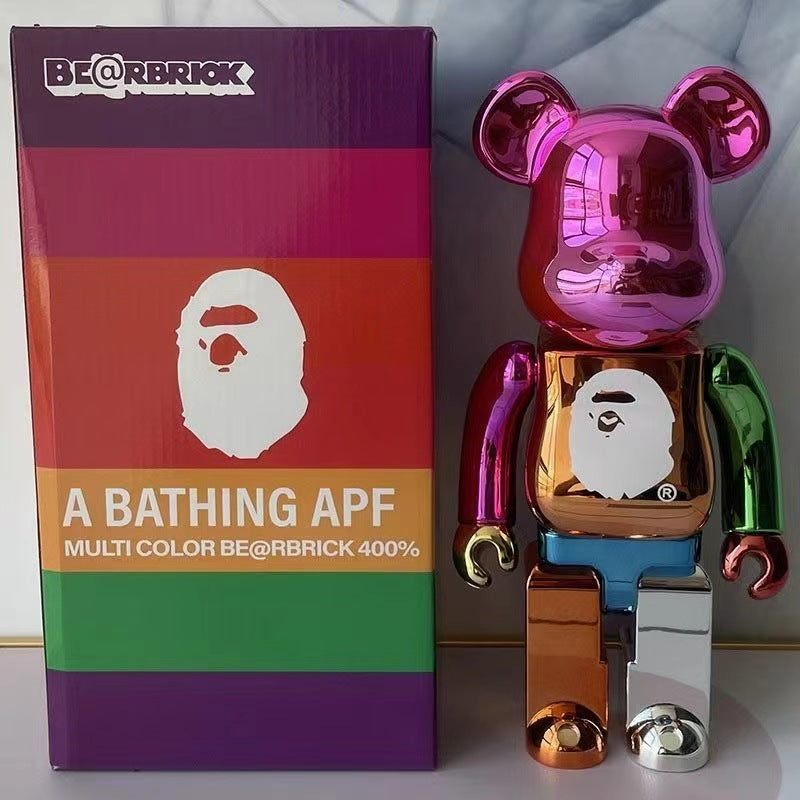 A metallic multi-color BE@RBRICK toy figure standing next to its branded retail box. The figure has a reflective chrome finish with pink, green, blue, orange, and silver sections, plus a white ape face graphic on the chest.
