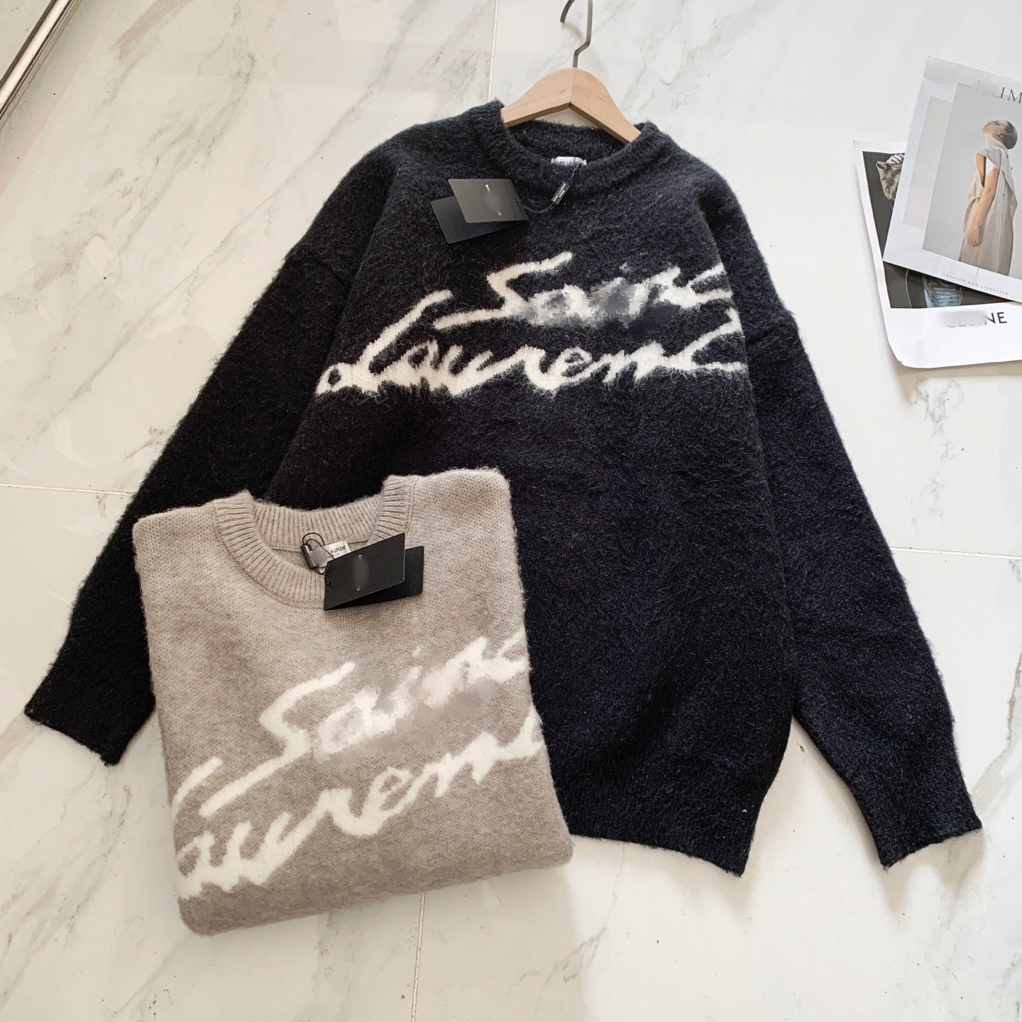 Two Saint Laurent fuzzy knit sweaters with white cursive logo lettering, displayed on a light marble surface. One sweater is dark navy blue hanging on a wooden hanger, and the other is folded in front in a light taupe-beige color.