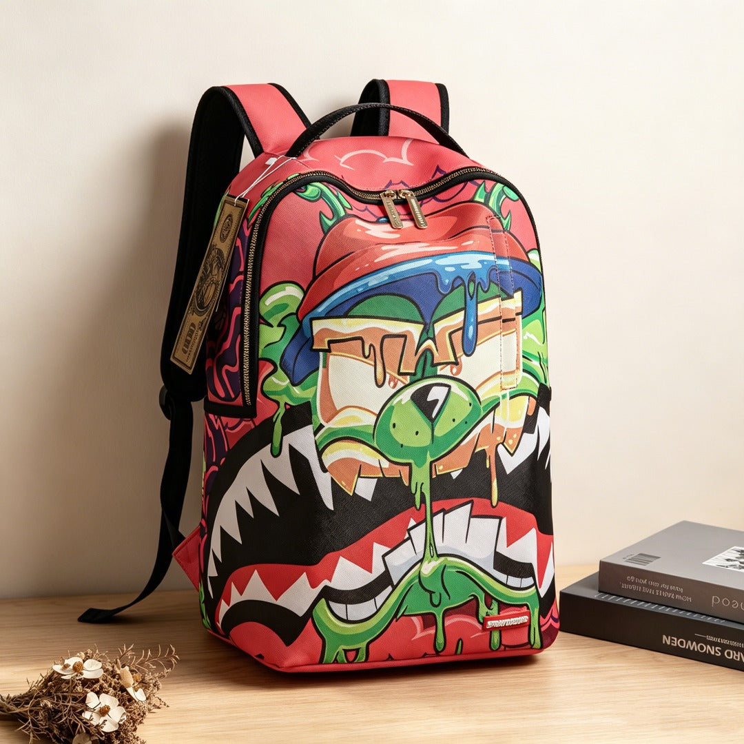 A red backpack with a colorful graffiti-style alien mouth graphic, shown on a wooden surface against a beige wall with a book and dried flowers nearby.