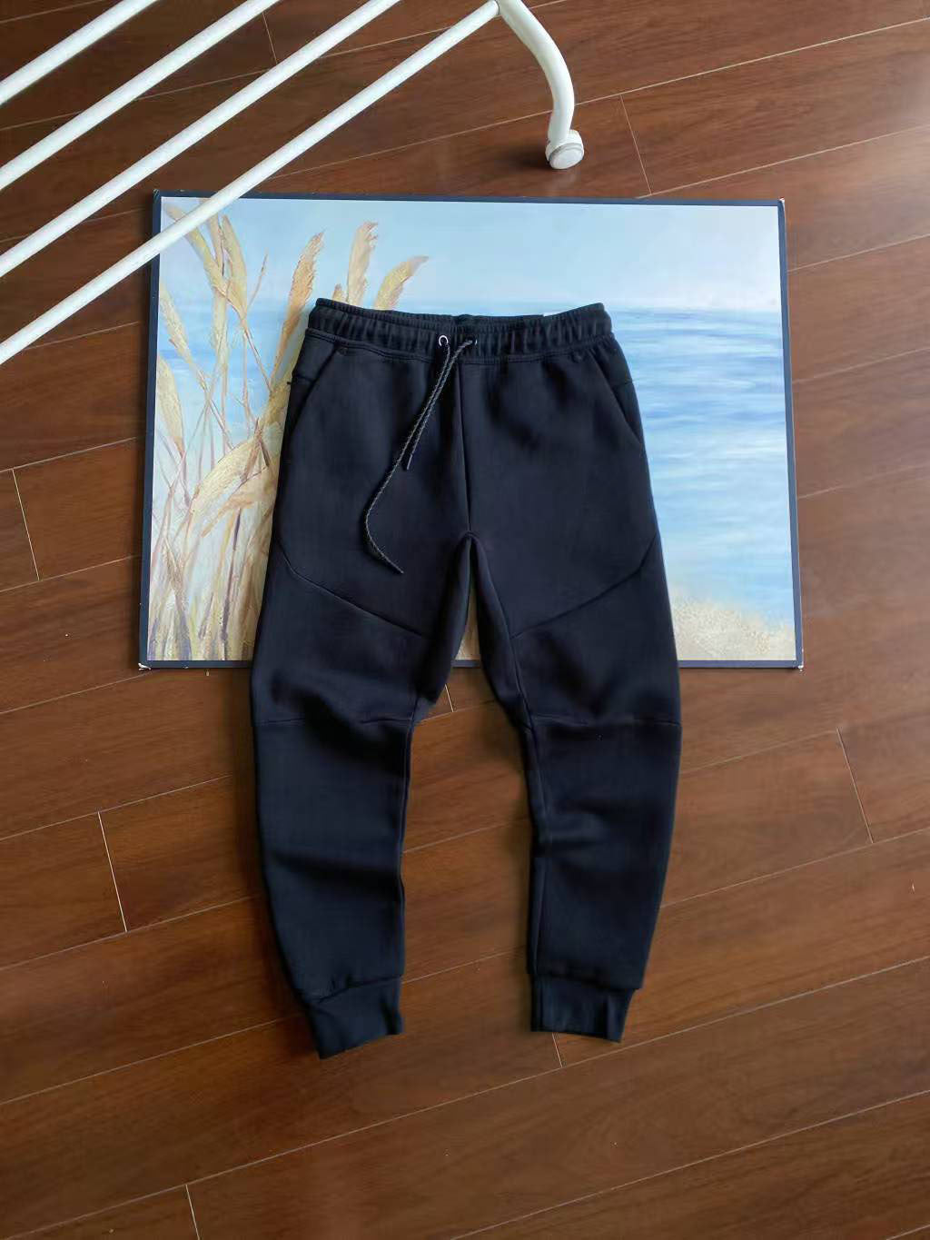Black jogger sweatpants with an elastic drawstring waist and tapered legs, laid flat on a wooden floor in front of a framed beach painting.