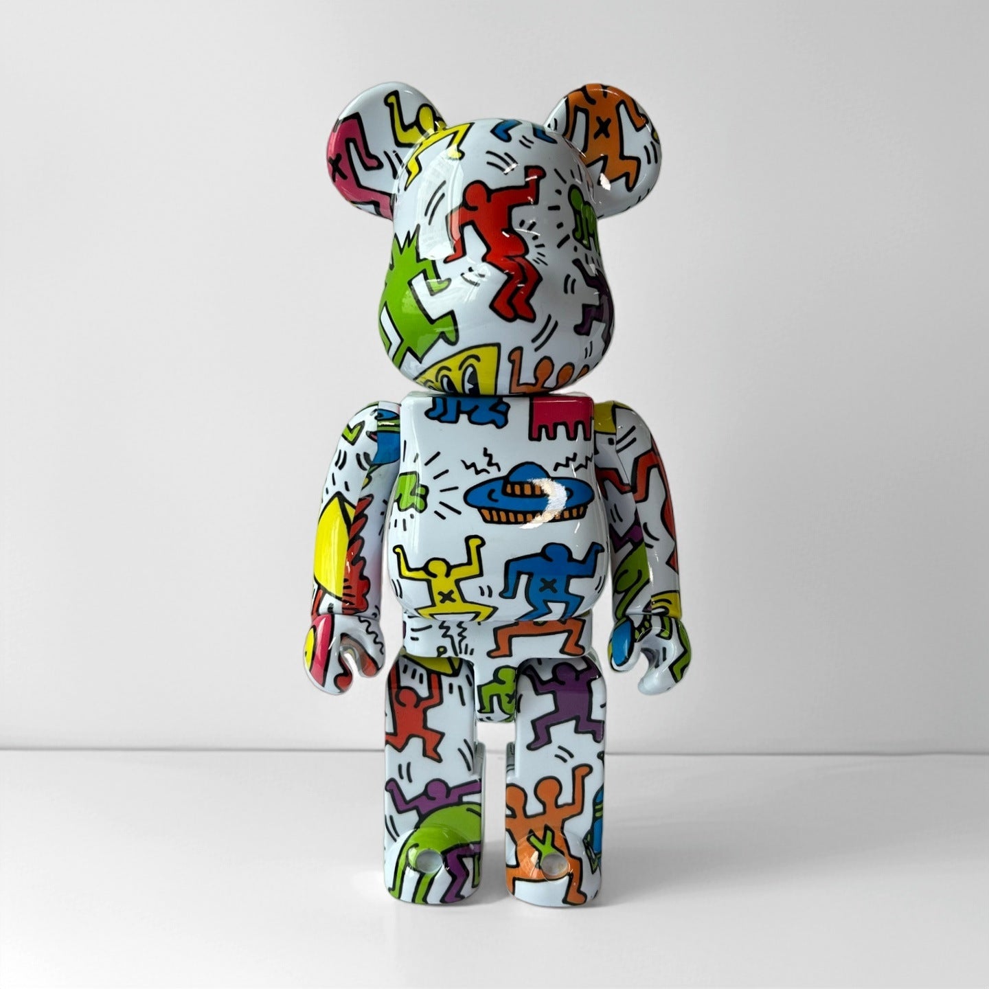 A collectible bear figure with a white base covered in colorful Keith Haring-style doodle patterns, photographed against a clean light gray studio background.