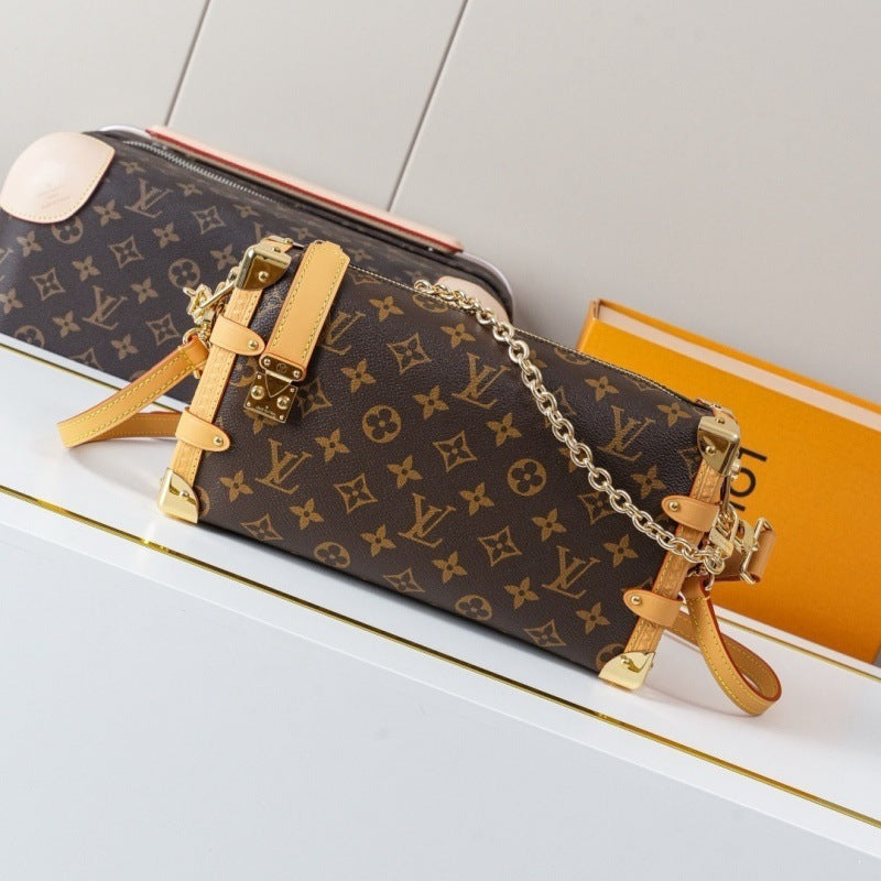 A Louis Vuitton monogram canvas shoulder bag with tan leather straps and gold-tone hardware, styled on a white shelf next to an orange book.