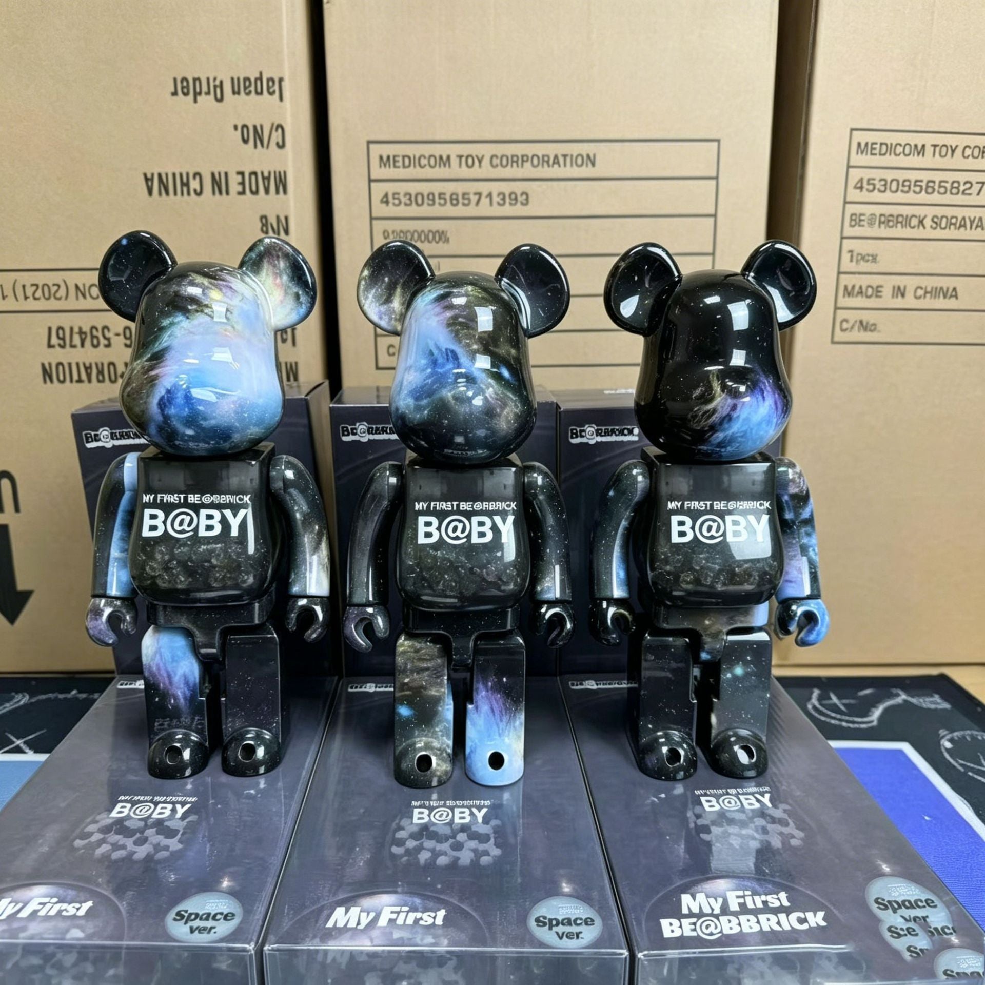 Three BE@BRICK Space War My First BE@BRICK figurines with a black and blue galaxy print, standing on their original packaging boxes in front of cardboard storage boxes.