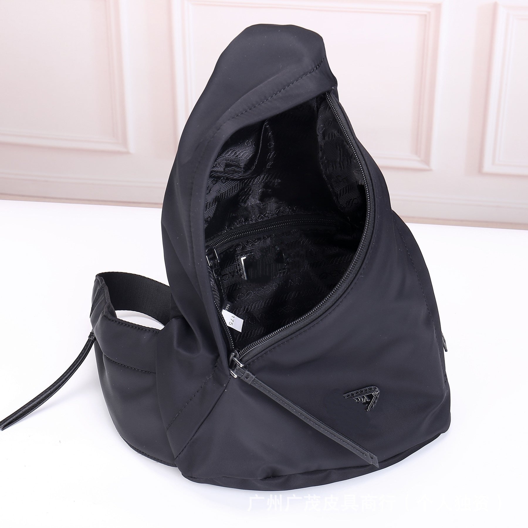 A black nylon sling backpack shown open to display its interior compartments and zipper details, set against a light pink paneled wall and white surface.