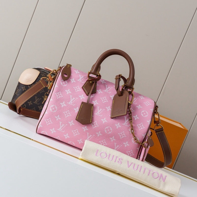 A pink Louis Vuitton Speedy handbag with brown leather trim and handles, displayed on a white shelf alongside a Louis Vuitton monogram pouch, an orange box, and a cream fabric bag tag printed with 'LOUIS VUITTON'.