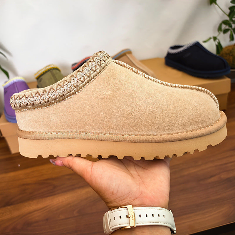 A beige suede slip-on clog-style shoe with braided trim and a chunky lug sole, held in a person's hand over a wooden surface. Other slippers are visible in the background.