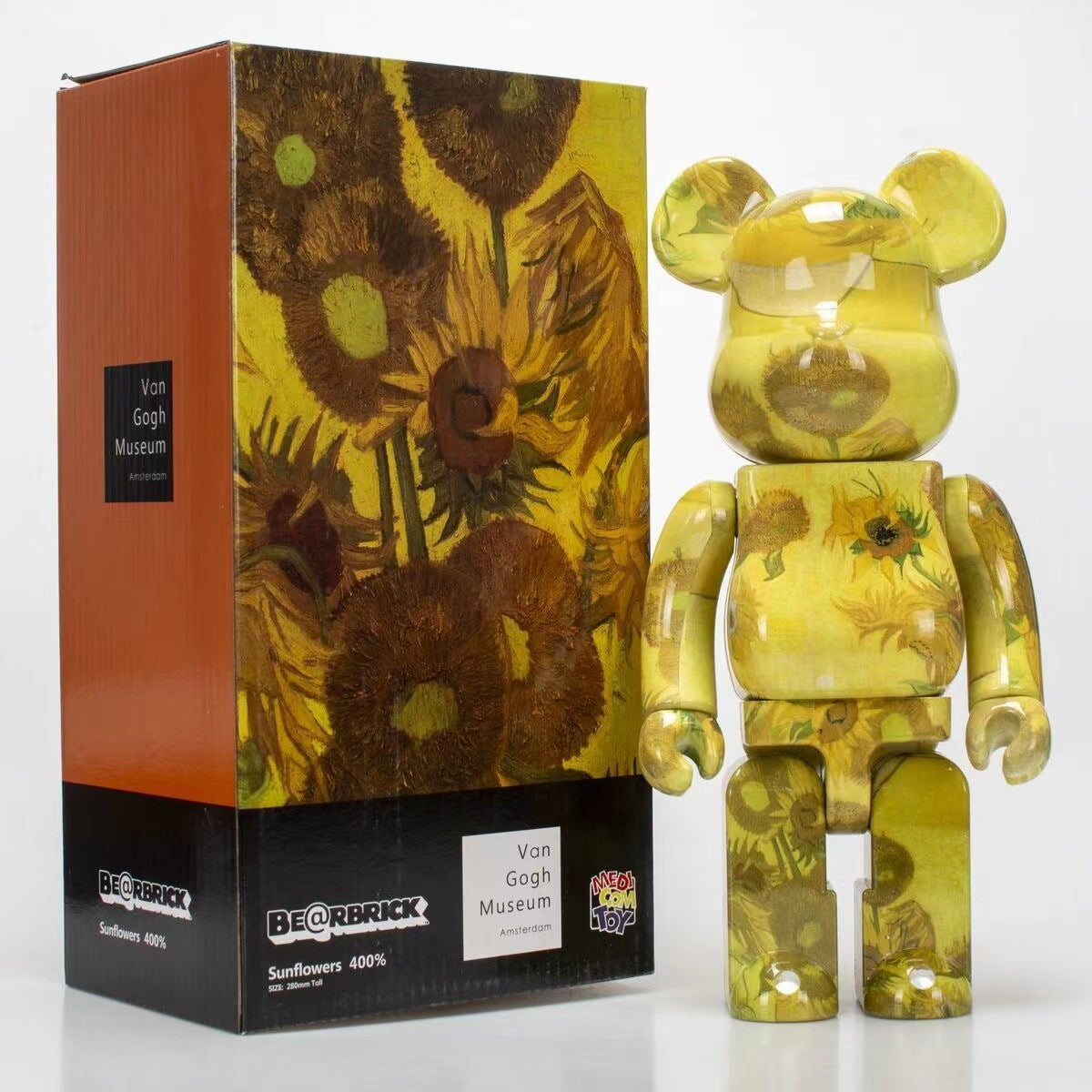 A translucent yellow Bearbrick collectible figure with a sunflower print stands next to its retail box. The packaging features a Van Gogh Museum design with a large sunflower artwork and a black base labeled 'Bearbrick' and 'Van Gogh Museum'.