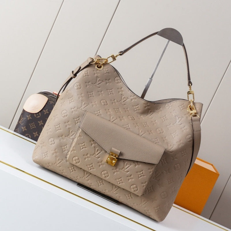 A beige Louis Vuitton monogram embossed hobo handbag with gold-tone hardware, displayed on a white shelf next to an orange box and a dark monogram pouch.