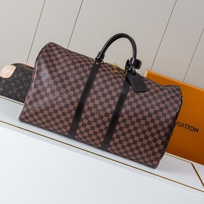 A Louis Vuitton duffle bag in brown and beige checkerboard canvas with black leather trim and handle, displayed on a white surface next to an orange Louis Vuitton box.