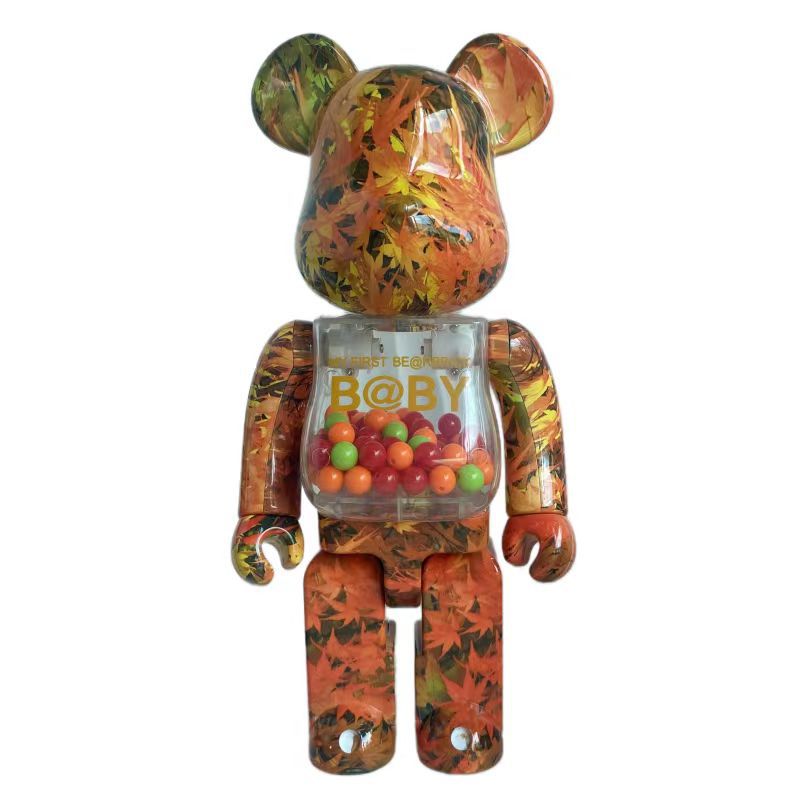 A Bearbrick toy figure with an autumn maple leaf pattern in orange, yellow, and green, featuring a clear chest compartment filled with colorful candies and 'B@BY' printed on the front, set against a plain white background.