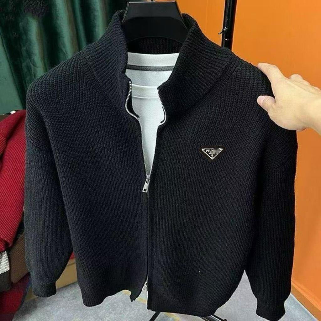 A black ribbed knit zip-up cardigan sweater with a stand collar and a small triangular metal logo on the chest, displayed on a hanger with a white shirt underneath. A hand is holding the right shoulder area.