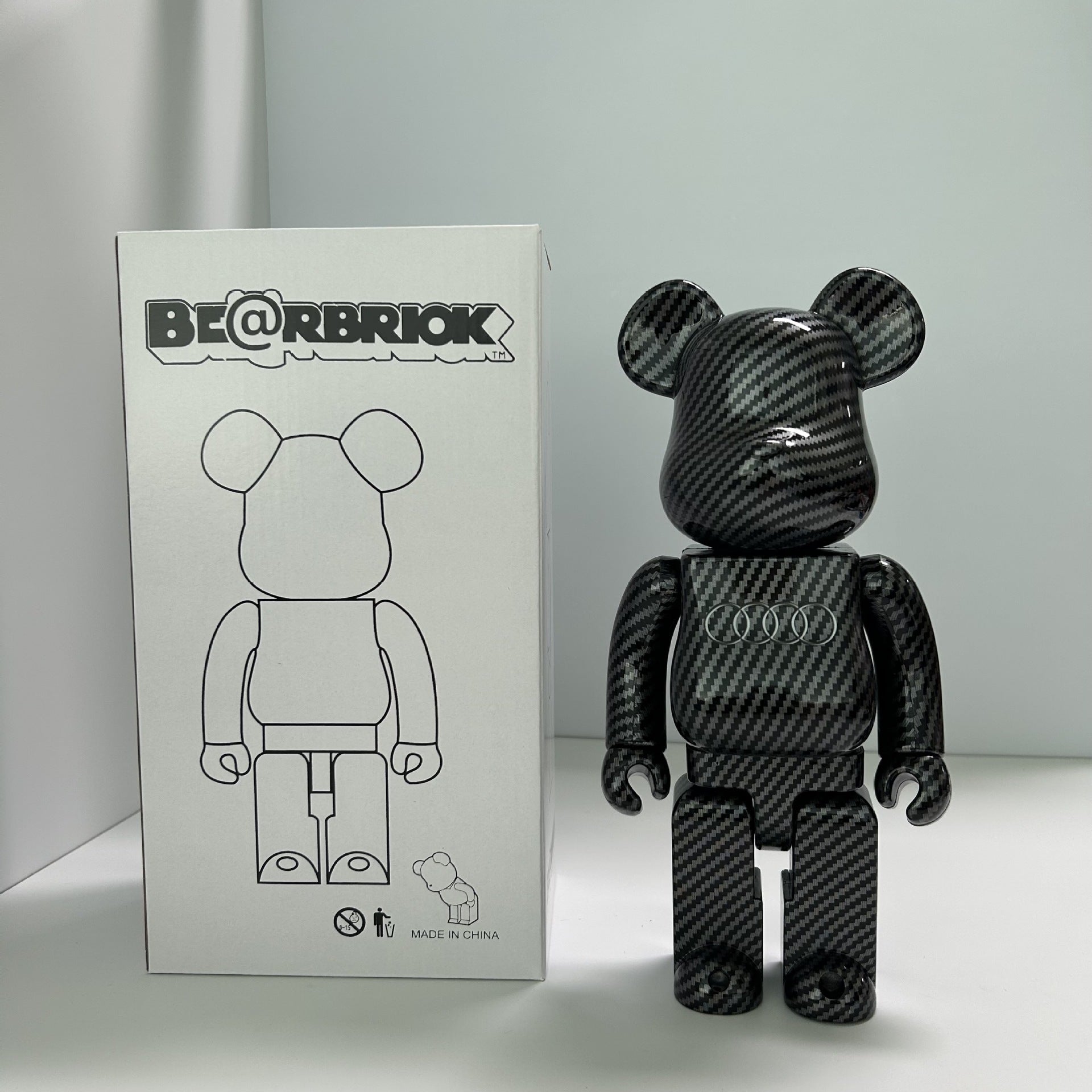 A Bearbrick collectible figure with a glossy black carbon fiber pattern stands next to its white packaging box, which features the BE@RBRIOK logo.