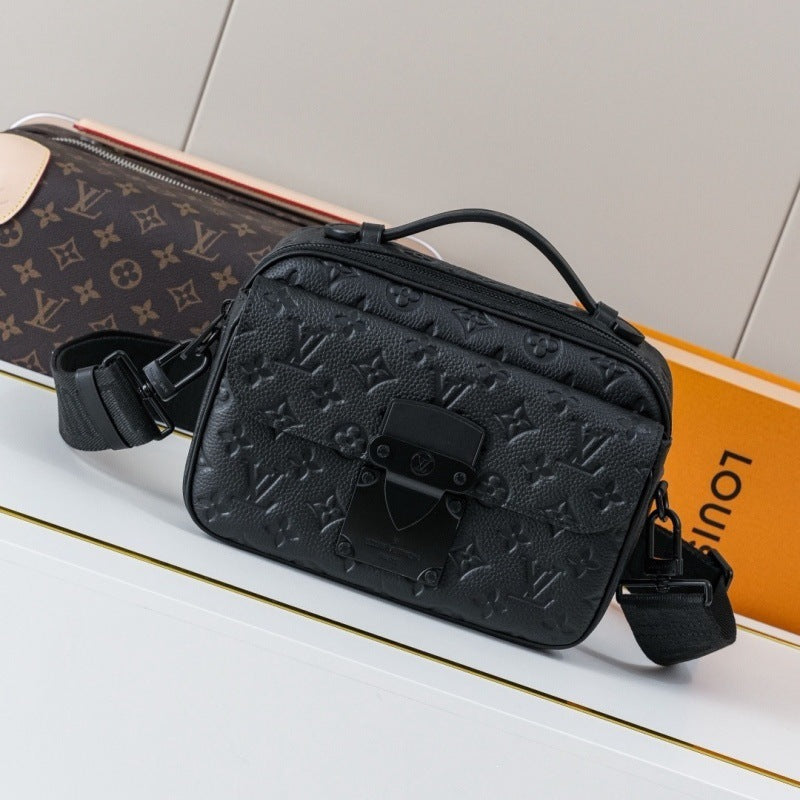 A black embossed leather crossbody bag with a top handle and adjustable shoulder strap, displayed on a white surface with a Louis Vuitton monogram pouch and orange box in the background.