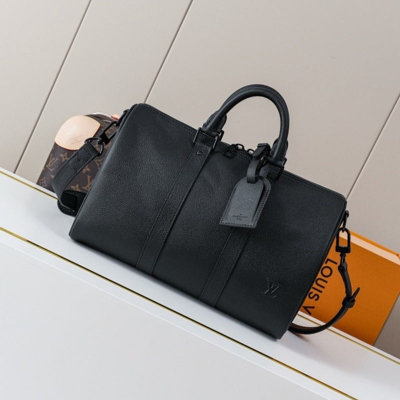 A black pebbled leather Louis Vuitton duffle bag with top handles and a detachable shoulder strap, displayed on a white shelf next to an orange Louis Vuitton box.