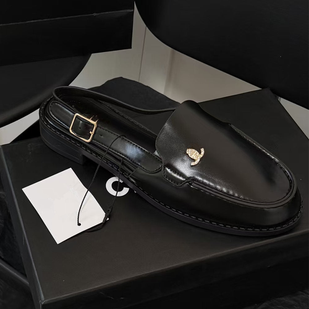 A black leather loafer shoe with a gold-tone logo charm, resting on a black shoebox with its white paper tag still attached.