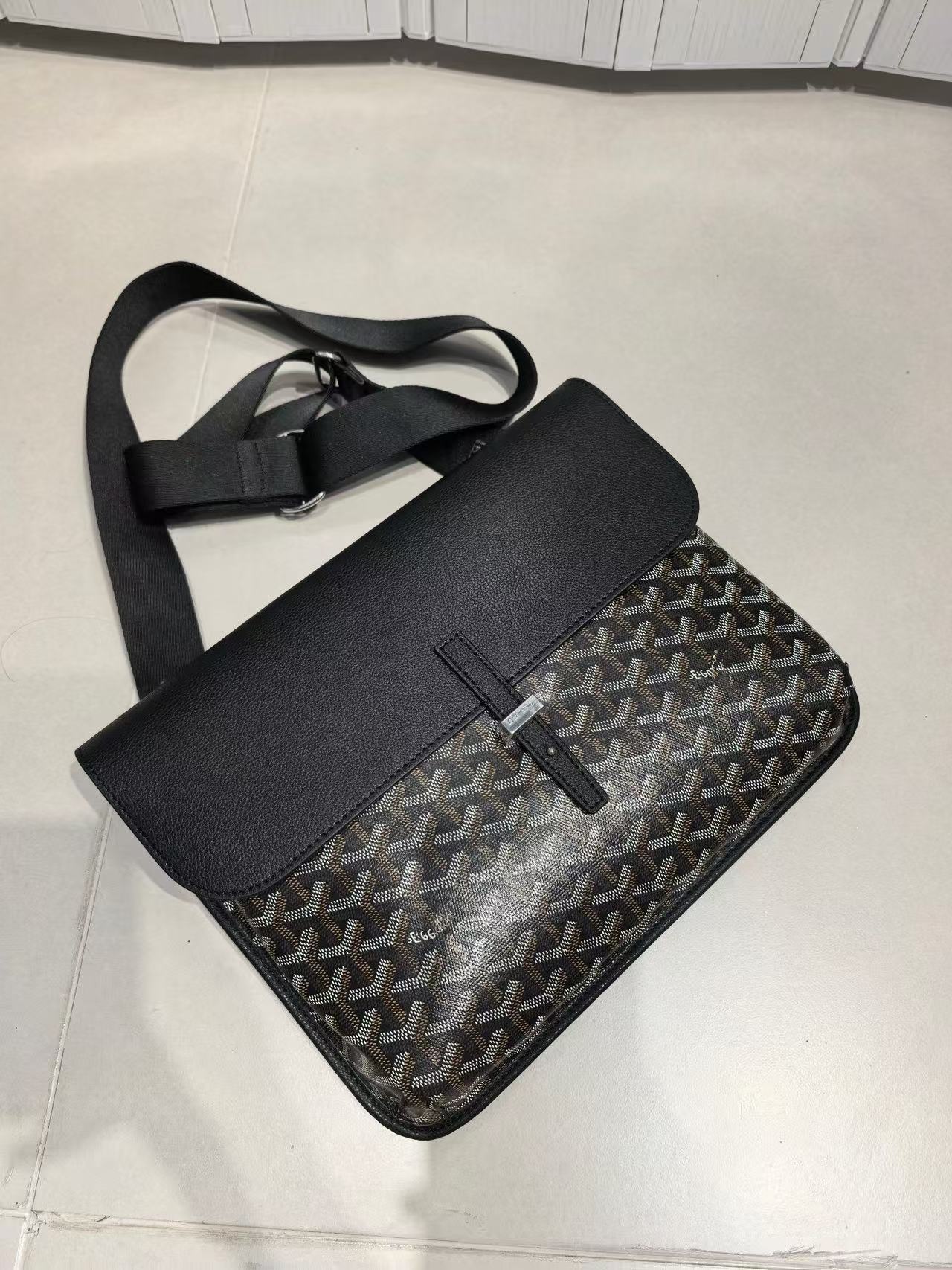 A Goyard messenger bag in black and gray Goyardine canvas with a black leather flap and adjustable shoulder strap, resting on a light-colored tiled floor.