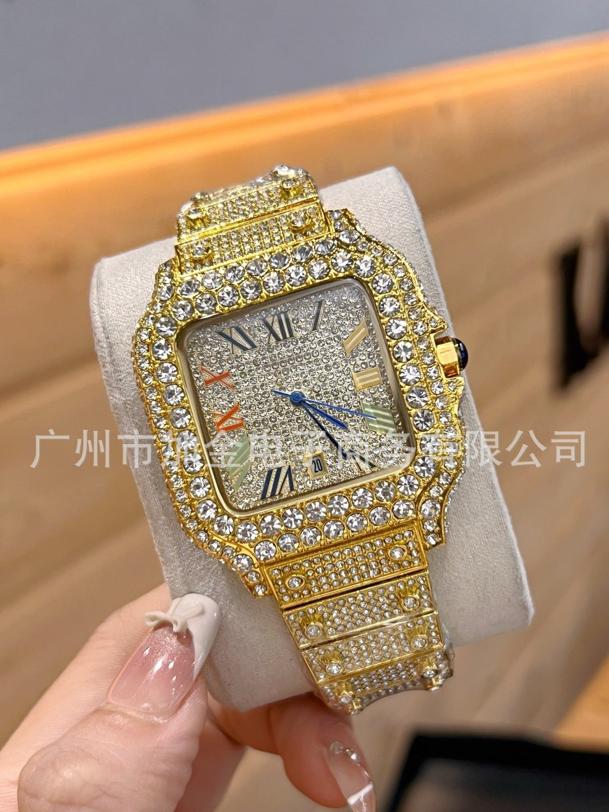 A gold-tone watch with a diamond-encrusted bezel and matching diamond-embellished bracelet, displayed on a light gray cushion and held by a hand. The watch face has a textured silver-gray background with Roman numerals and small orange accents.