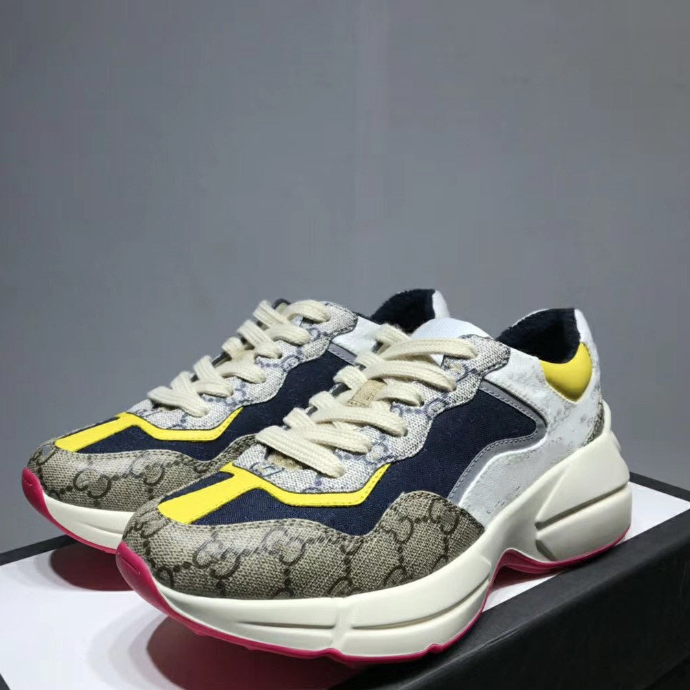A pair of low-top sneakers with a beige and gray monogram pattern, navy blue overlays, cream laces, a bright yellow accent stripe, and a chunky cream midsole with a pink outsole, displayed on a black platform against a gray background.