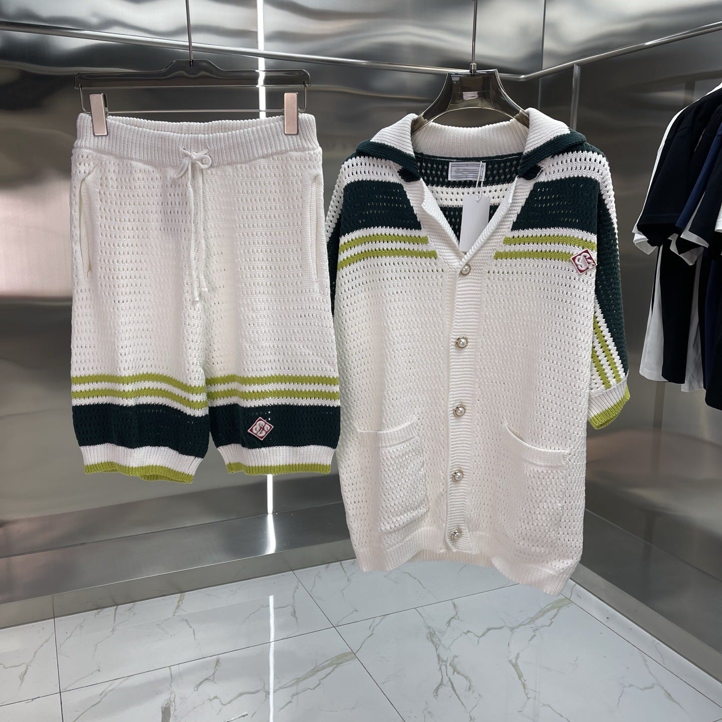 A matching knit lounge set hanging on a rack, featuring white textured shorts with dark green and olive green stripes, and a white hooded cardigan with dark green and olive green stripes on the sleeves and shoulders.