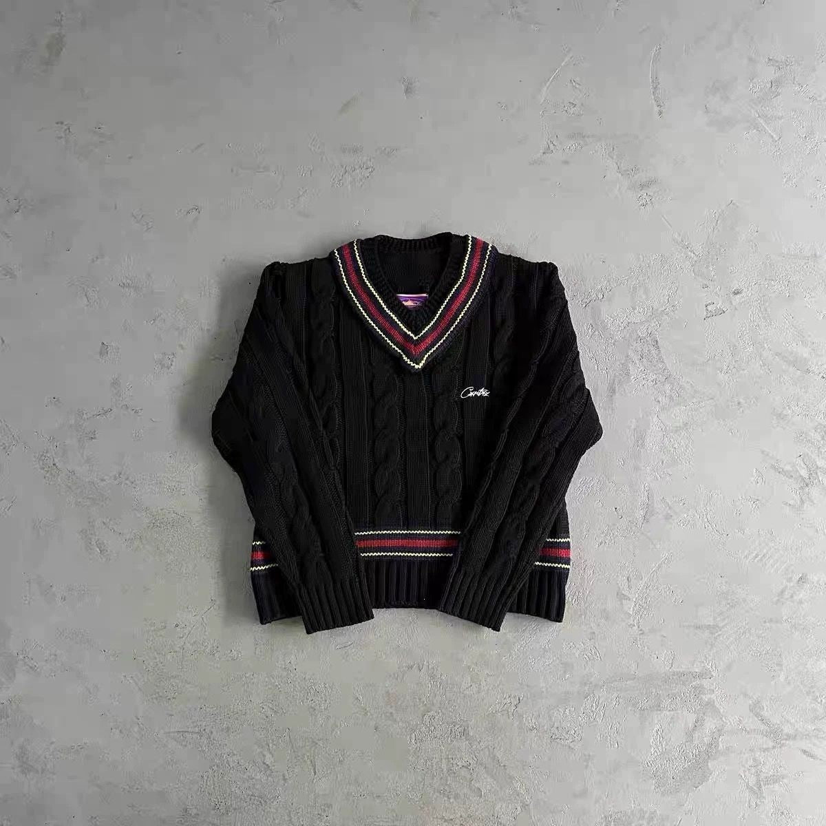 A black cable-knit V-neck sweater with red, white, and green striped ribbed trim on the collar and cuffs, laid flat on a textured gray concrete surface.