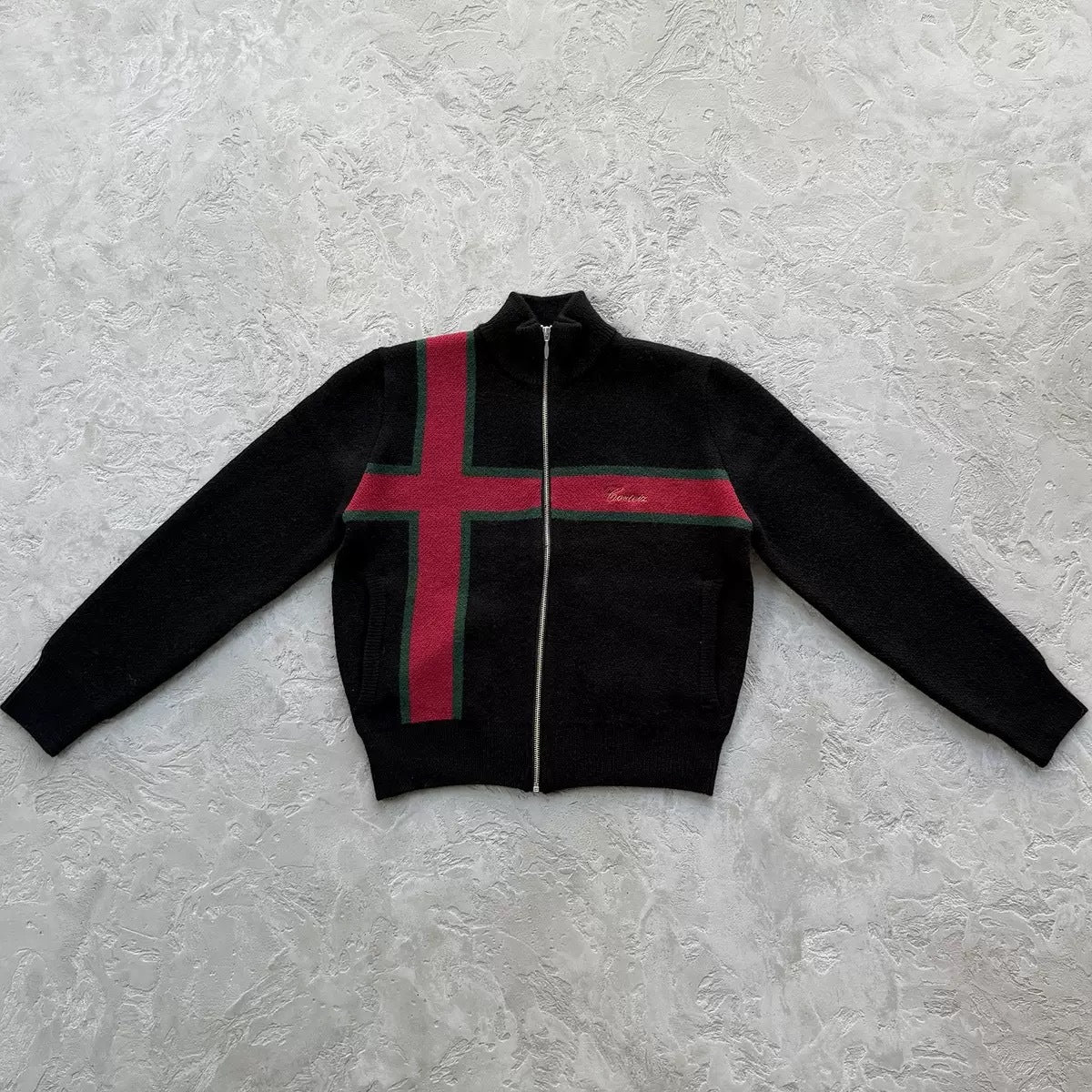 A black zip-up knit sweater with a red and green striped cross design across the chest, laid flat on a textured light gray surface.