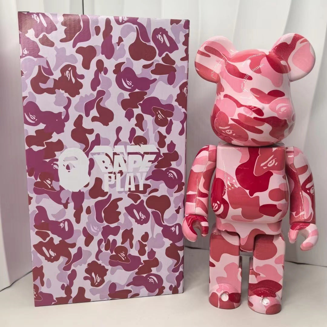 A pink camouflage Bearbrick toy figure standing next to its branded packaging box, which features a matching pink and purple camo pattern with a white 'BAPE PLAY' logo.