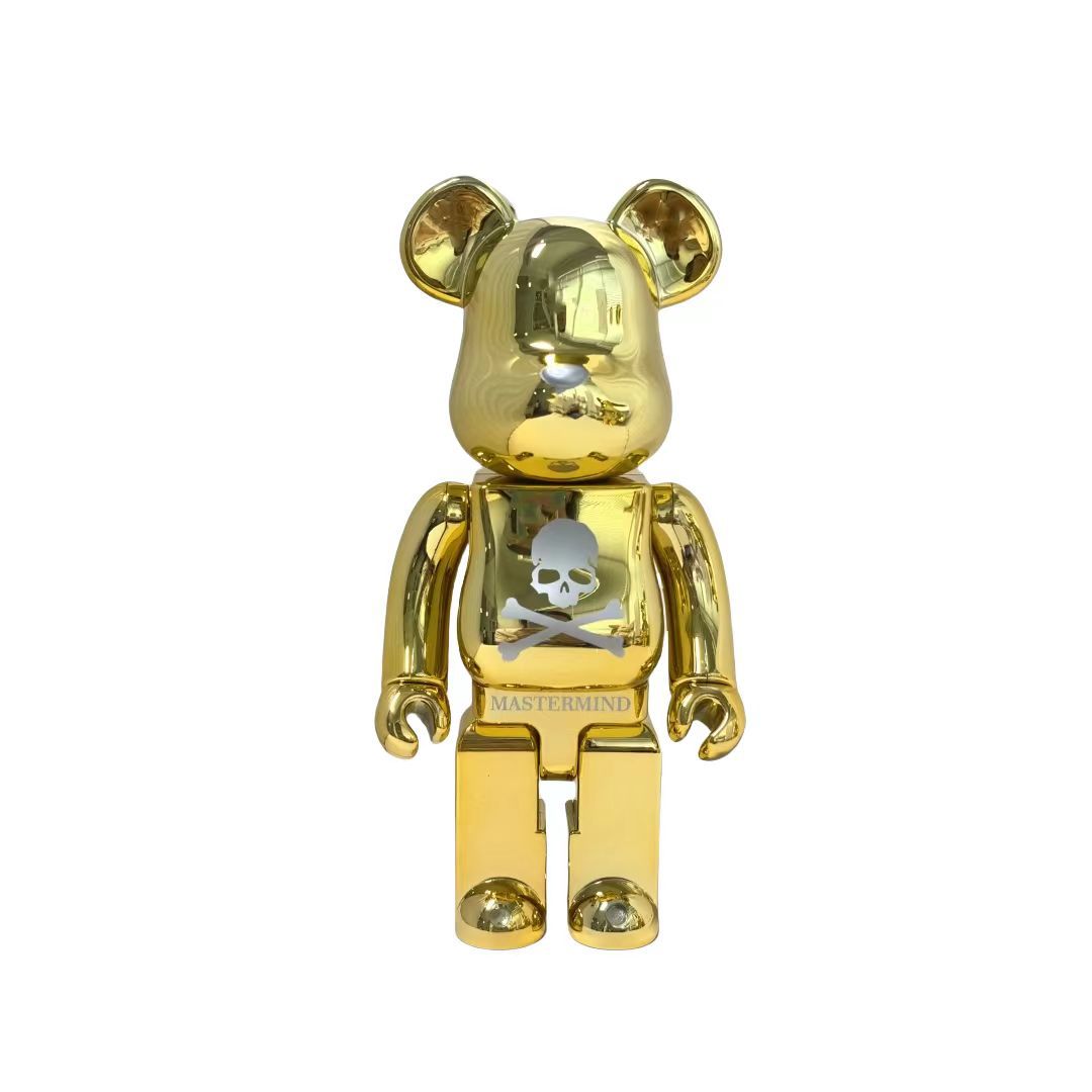 A metallic gold Bearbrick figure with a skull and crossbones graphic on the chest and the text 'MASTERMIND' on the lower torso, shown against a plain white background.