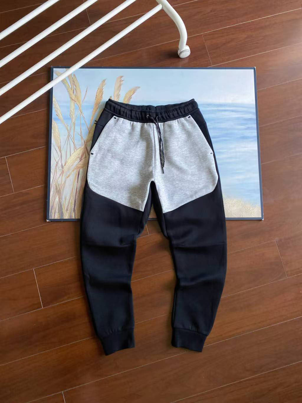 A pair of color-block jogger sweatpants with a light gray and black design, laid flat on a wooden floor in front of a framed beach painting.