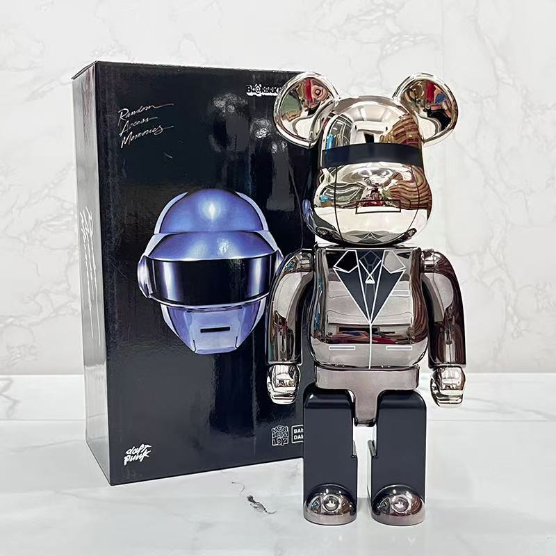 A metallic bear-shaped collectible figure standing in front of its black retail box. The figure has a chrome silver finish with a black visor and suit details, and the box features a blue helmet graphic and stylized script text.