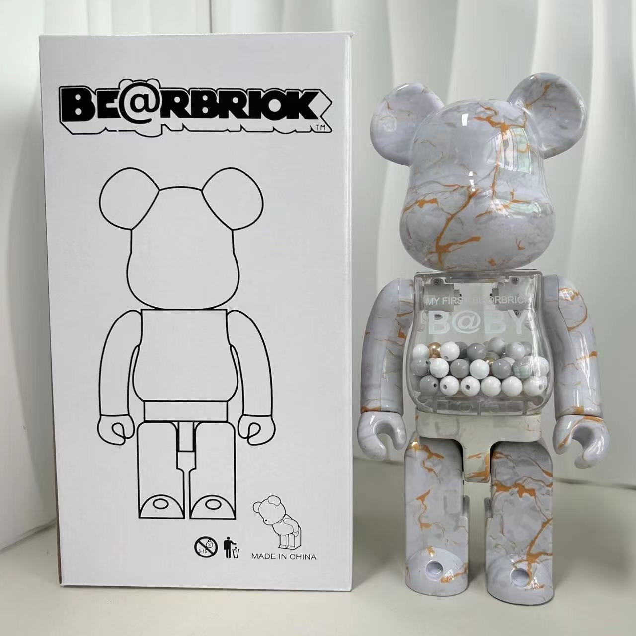 A Bearbrick collectible figure with a white and gray marble pattern and gold veins, standing next to its white packaging box. The box features the BE@RBRIOK brand logo and a black line drawing of the bear figure.