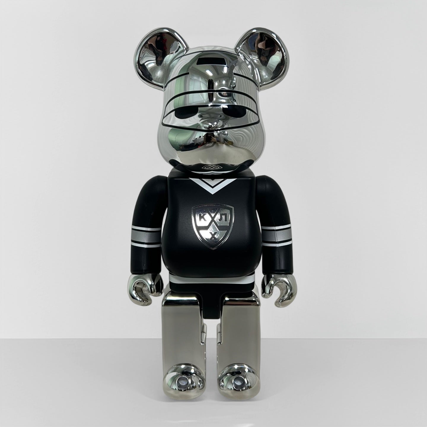 A metallic bear-shaped collectible figure standing upright against a plain light gray studio background. The figure has a reflective silver chrome finish on its head, ears, and legs, with a black body and a silver-and-white athletic-style crest on the chest.