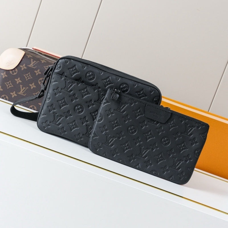 A black embossed monogram wallet and matching pouch displayed on a white surface, with a brown monogram bag partially visible behind them against a light neutral background.