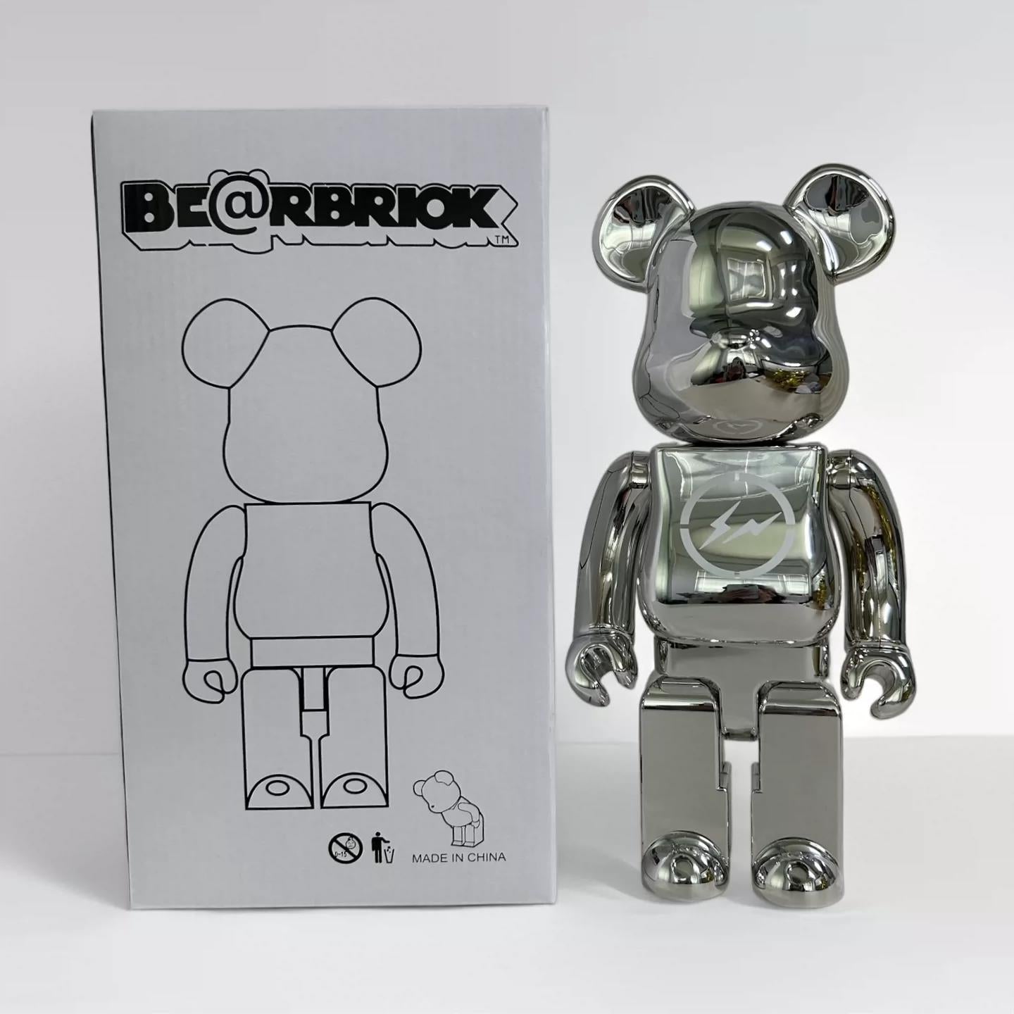 A metallic silver Bearbrick toy figure standing next to its original white packaging box, which features the @RBRIOK logo and a black line drawing of the bear figure.