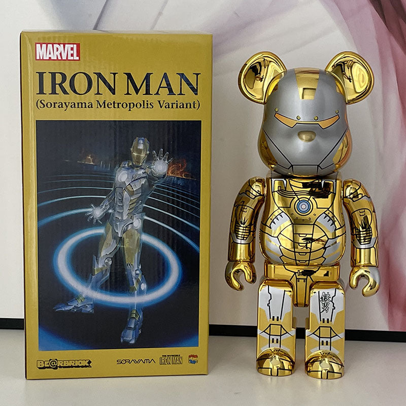 A shiny gold and silver Iron Man Sorayama Metropolis Variant bear figure stands next to its original yellow and black packaging box on a white surface.