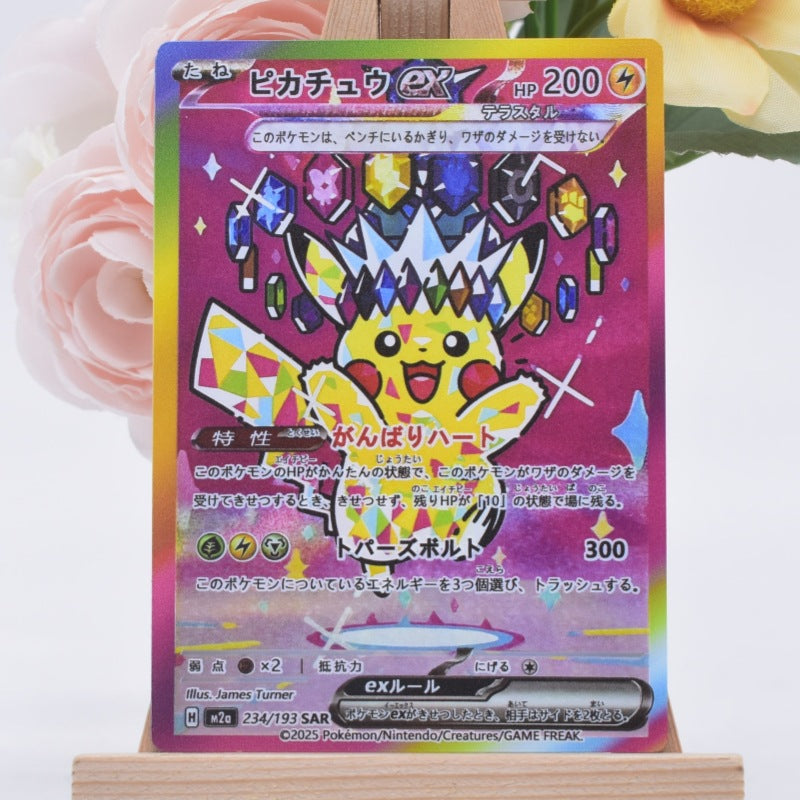 A Pokémon trading card featuring Pikachu with a colorful gem crown, photographed upright on a small wooden display stand against a soft white background with blurred pink flowers.