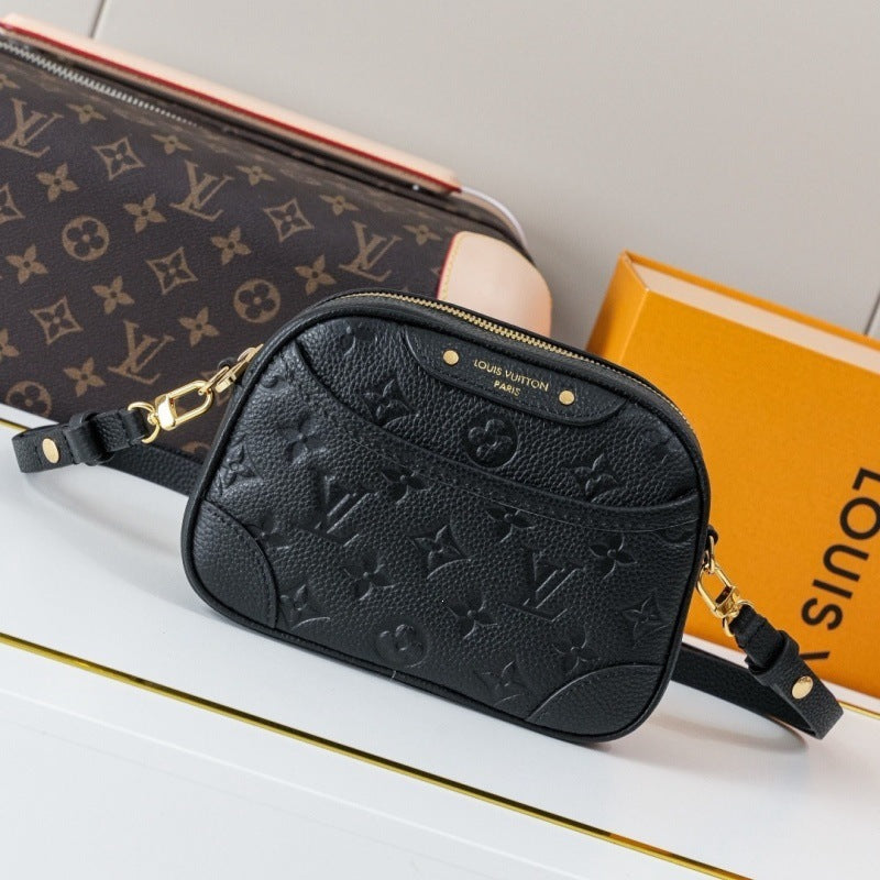 A black Louis Vuitton monogram embossed leather camera bag with gold-tone hardware, displayed on a white surface with a Louis Vuitton Speedy case and orange box in the background.