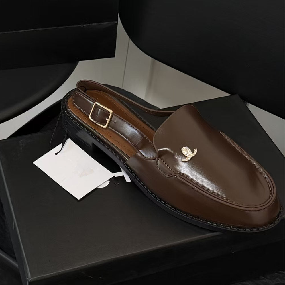 A brown leather slip-on loafer-style shoe with a small gold logo detail on the toe, displayed on a black shoebox with a white paper tag attached.