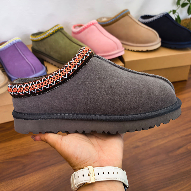 A hand holds a dark gray suede slipper with a colorful geometric embroidered trim. In the background, several other suede slippers in purple, green, pink, tan, and navy are displayed on wooden boxes on a wooden floor.