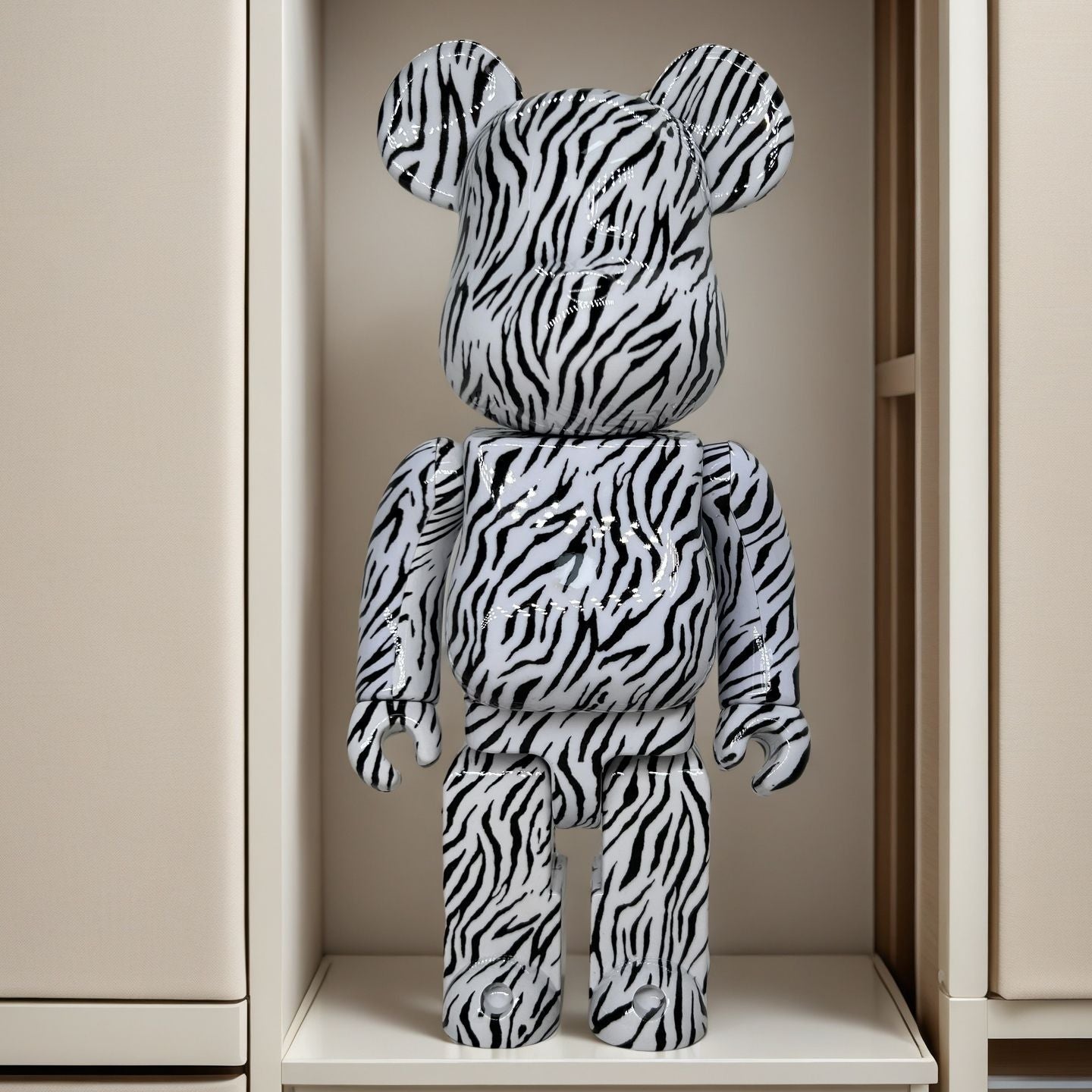 A bear-shaped collectible figure with a black and white zebra print pattern, standing on a shelf inside a beige cabinet or display case.