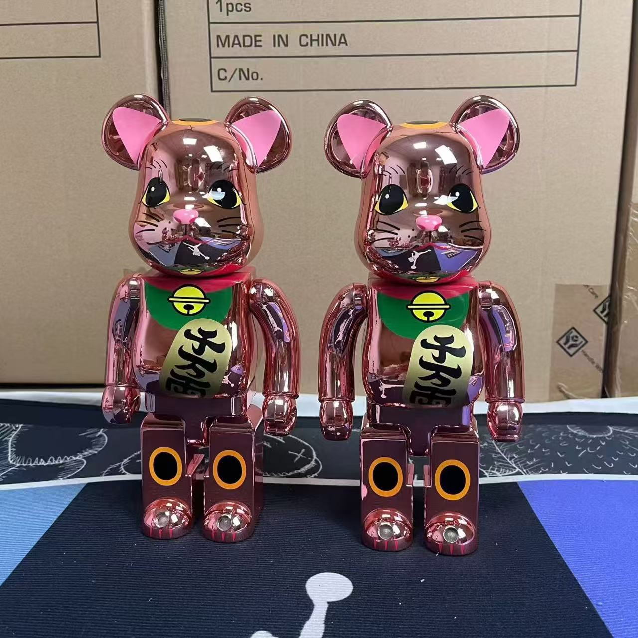 Two metallic rose gold bear figurines with pink ears and green bibs, standing in front of a cardboard shipping box.